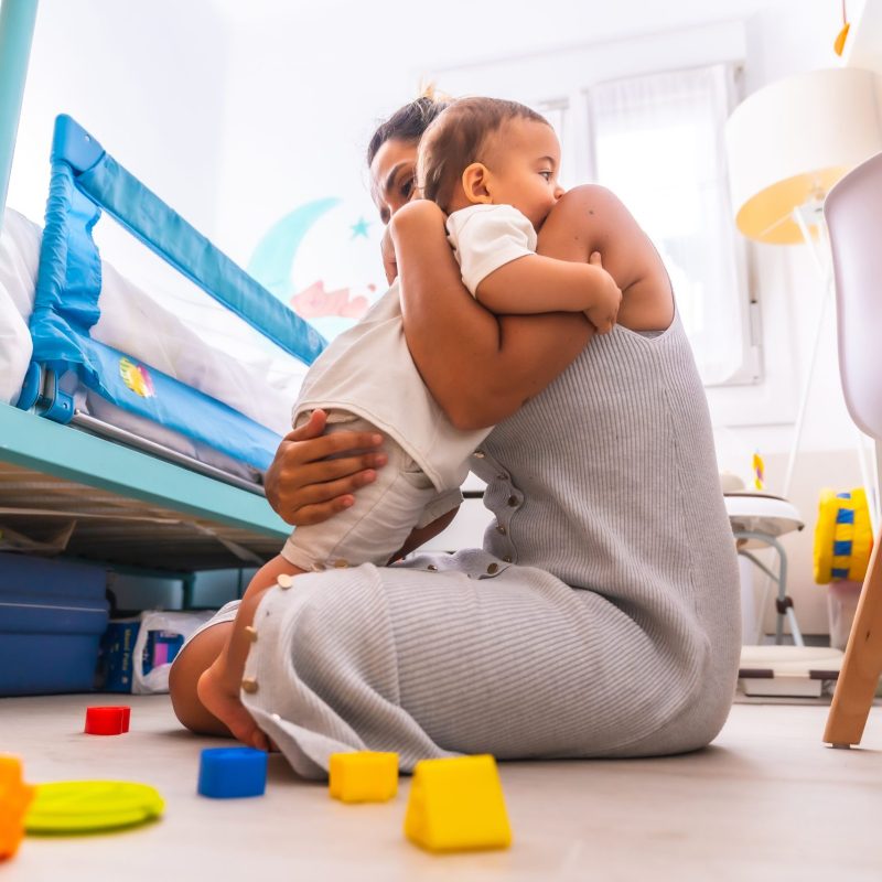 Young Caucasian mother playing with her in the room with toys. Baby less than a year learning the first lessons of her mother. Mother playing with her son and hugging him lovingly